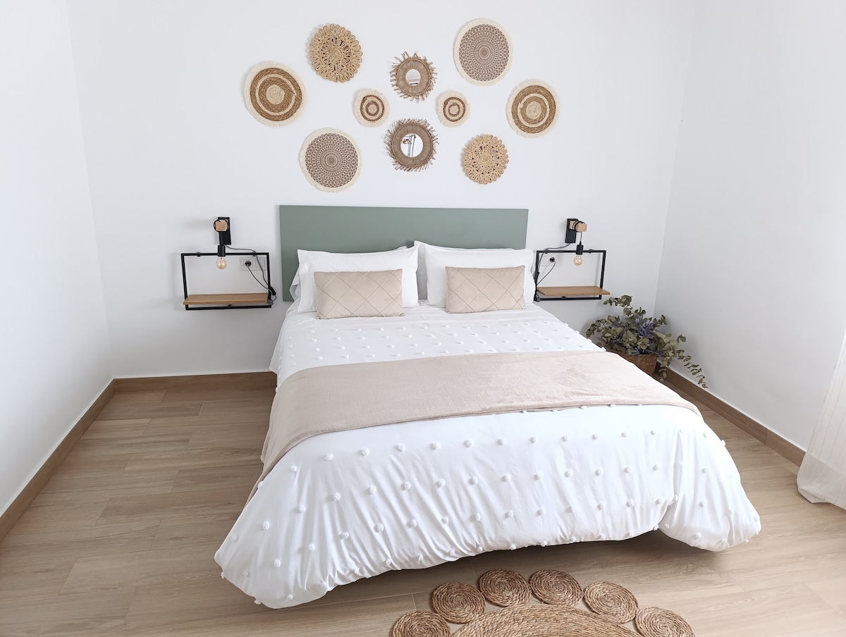 A serene bedroom displays a spacious bed with textured bedding and neutral tones. Two bedside tables with lamps are positioned on either side. Decorative wall art made of woven materials is arranged above the bed, adding a touch of warmth to the minimalist space.