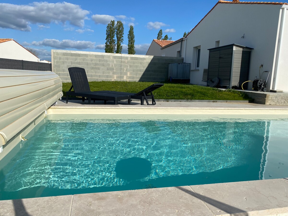 A clear pool is framed by a light-colored stone border, with a single black lounge chair positioned at the poolside. The surrounding area features green grass and a gray wall in the background, with houses visible nearby under a partly cloudy sky.