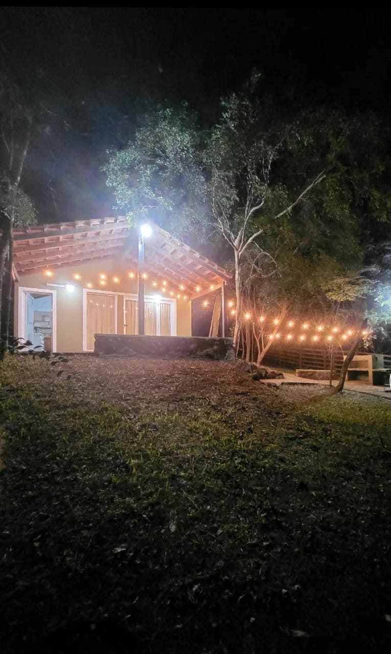 A charming cabin is set against a backdrop of natural greenery, illuminated by warm string lights. The structure features a sloped roof and multiple doors, creating an inviting outdoor space that encourages connection with nature.
