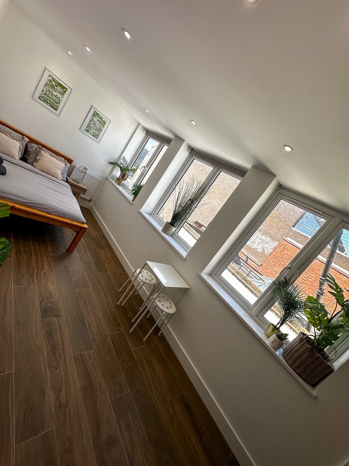 A bright and airy bedroom features a spacious bed complemented by decorative pillows. Large windows allow natural light to fill the room, showcasing modern window treatments. Potted plants are positioned in the corners, adding a touch of greenery, while a small seating area is visible near the bed.