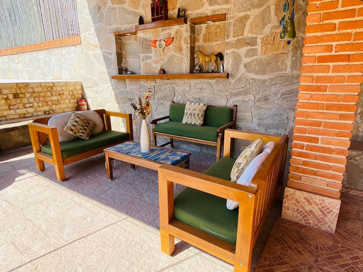 An outdoor seating area is featured, surrounded by stone walls. Two wooden couches and a coffee table provide a cozy space. Decorative items and a dried floral arrangement are placed on the table. Sunlight softly illuminates the area, creating a relaxing atmosphere.