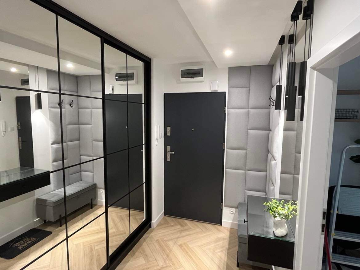 A modern entryway is presented, featuring a sleek black door and a grid of reflective panels that enhance the sense of space. A stylish bench is situated near the door, and subtle lighting is provided by hanging fixtures above. A small decorative plant adds a touch of greenery.