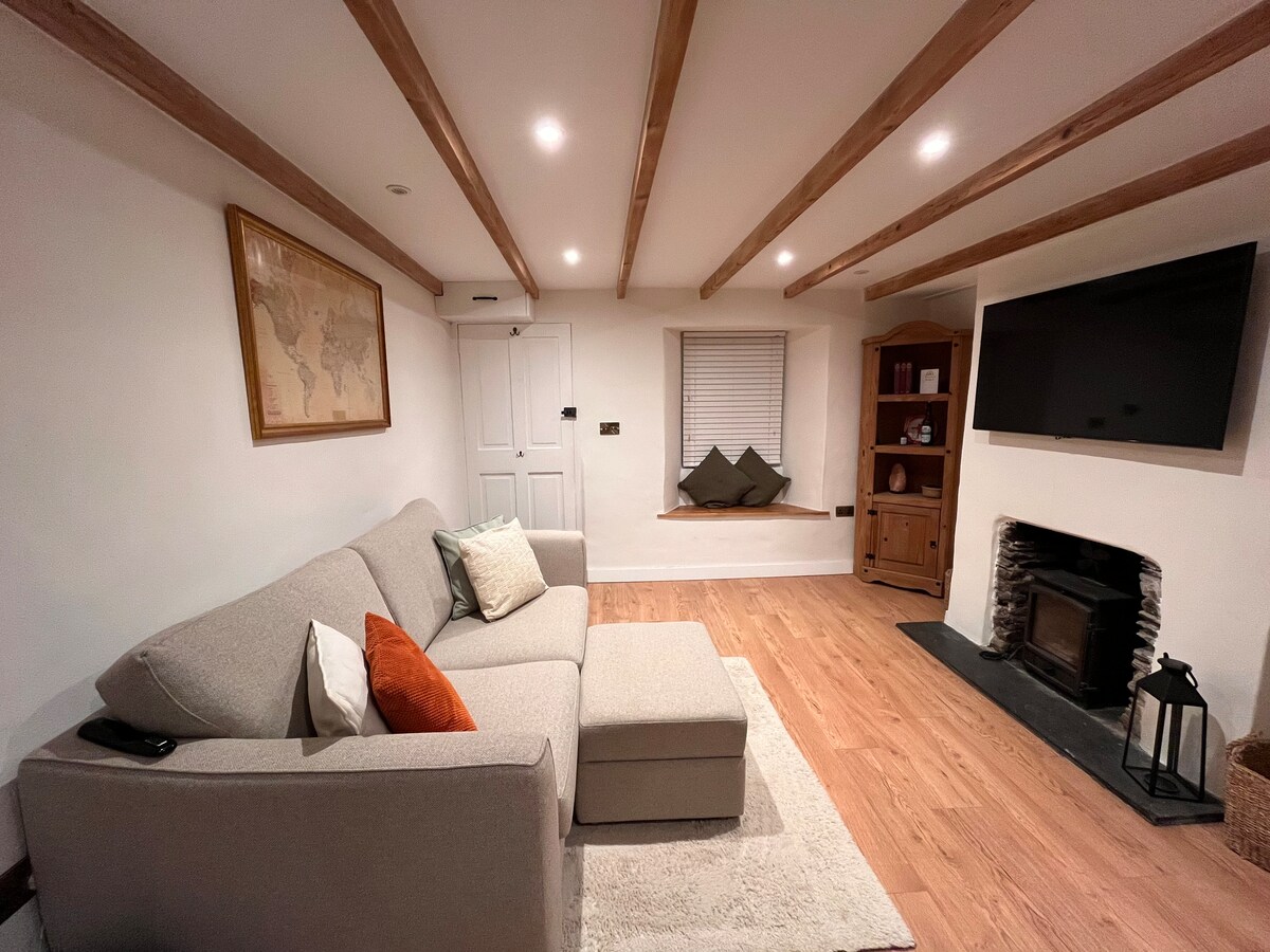 A cozy lounge area features a comfortable sofa adorned with decorative cushions. A smart TV is mounted on the wall opposite a log burner, with a charming window seat designed for relaxation. Warm wood beams accentuate the ceiling, enhancing the inviting atmosphere.