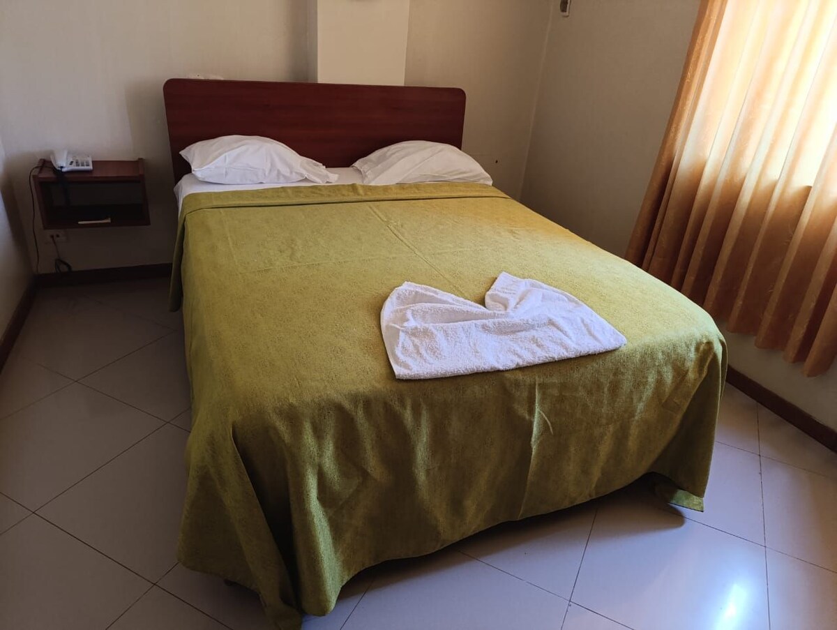 A neatly made bed is covered with a green spread and has white pillows arranged at the headboard. A towel is folded into a shape on one side. Natural light pours through a window covered with soft, sheer curtains.