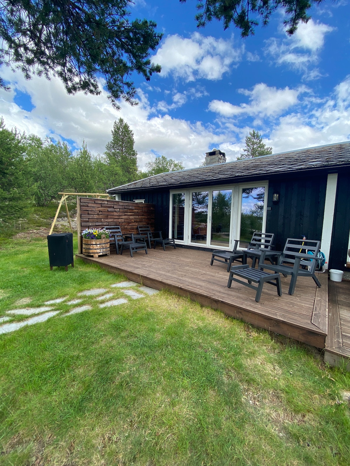 A spacious wooden deck area features several black outdoor chairs arranged for relaxation. A small flower arrangement is visible in a planter beside the entrance. The backdrop showcases a lush green landscape under a partly cloudy sky.