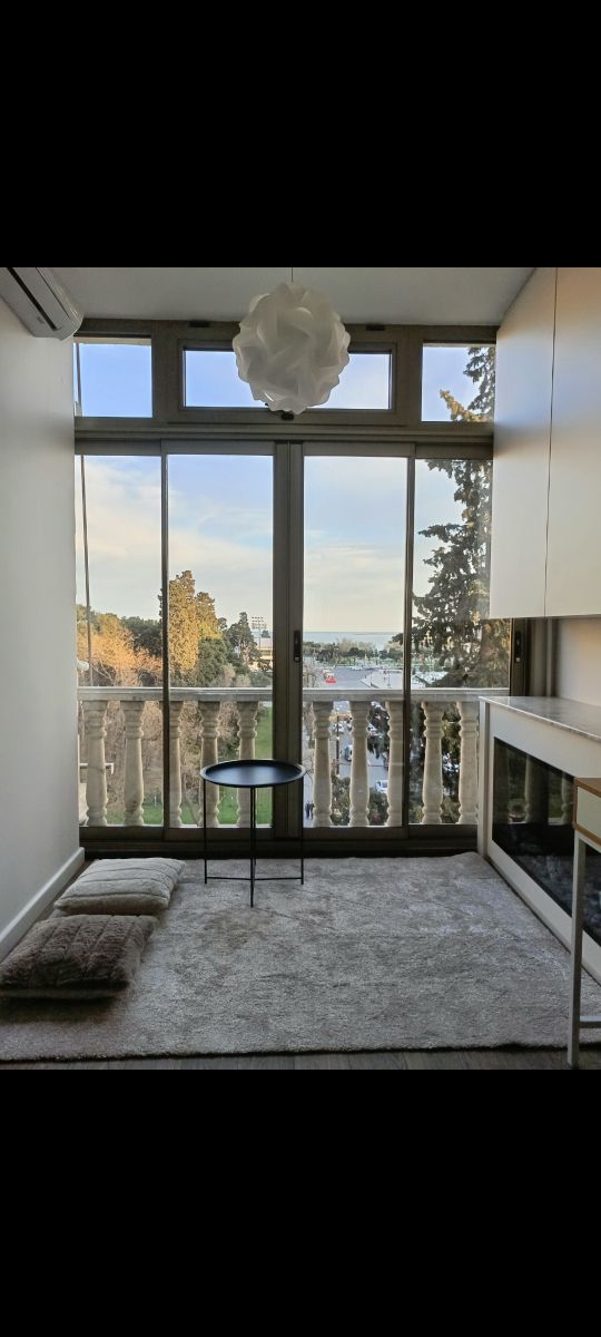 A bright sitting area features large glass doors that open to a scenic view, allowing natural light to fill the space. A small round table is positioned on a soft carpet, complemented by a cushion nearby. Greenery is visible outside, enhancing the serene atmosphere.