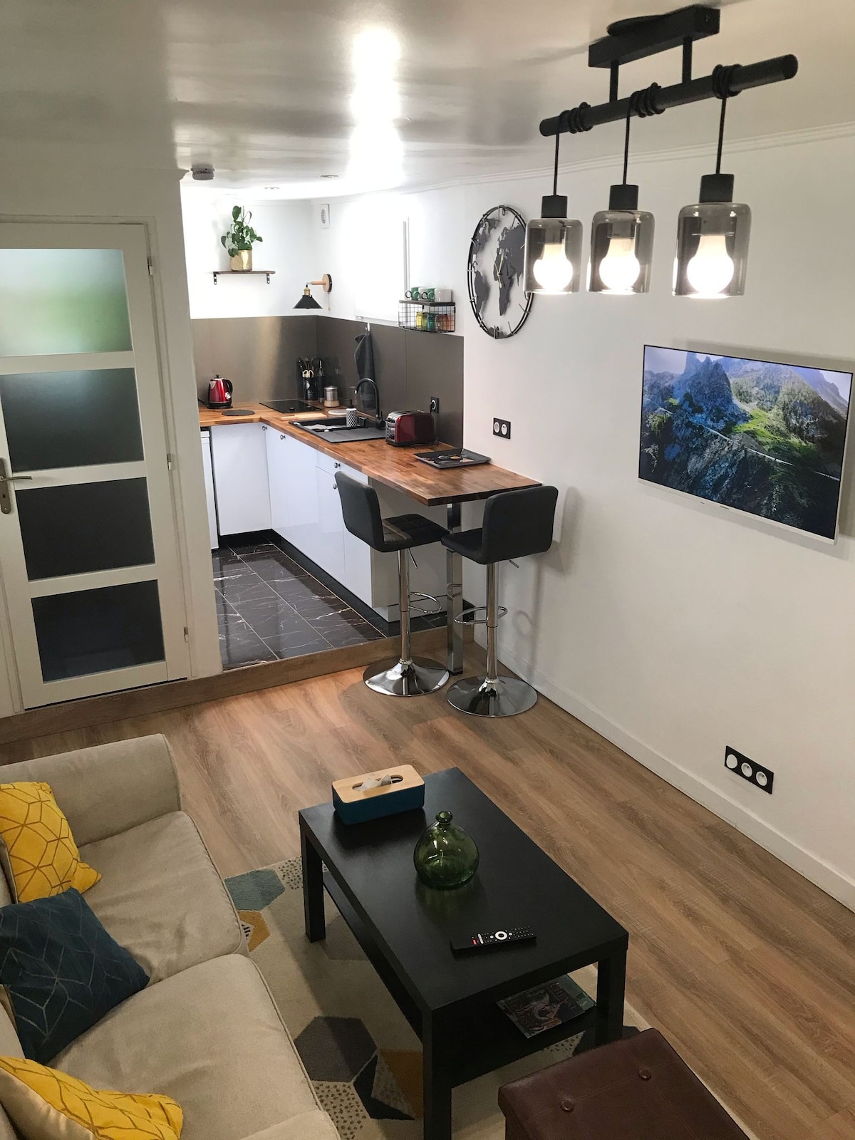 The studio presents a combined living and kitchen area. A compact kitchenette features modern appliances and counter space. Stylish bar stools are positioned at the counter, and a comfortable sofa sits nearby. Natural light enters through a window, highlighting the clean and functional design.