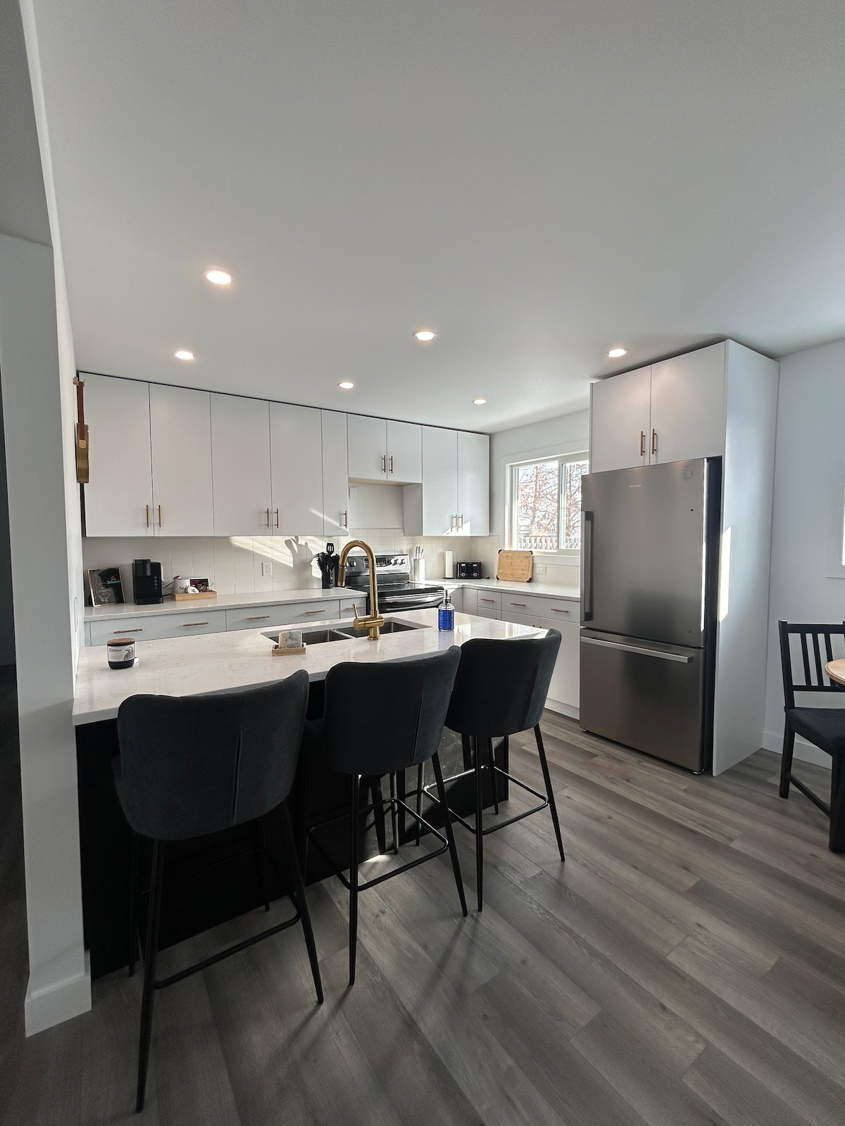 The kitchen showcases modern cabinetry in white and dark tones, complemented by stainless steel appliances. A spacious island with seating for four is positioned in the foreground, while natural light enters through a large window, illuminating the sleek countertops.