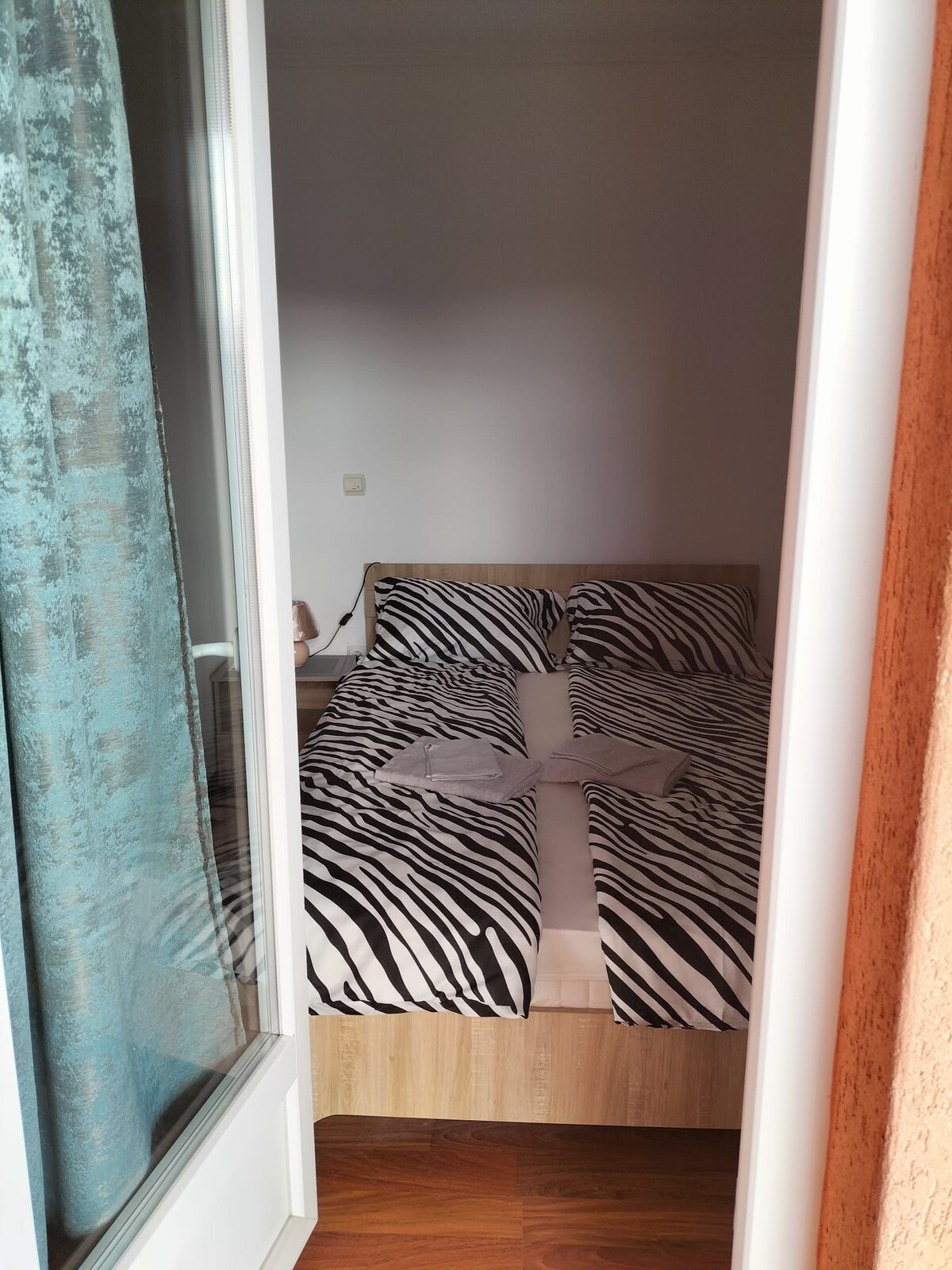 A cozy bedroom is visible through an open door. The space features a light wooden bed with a zebra-striped bedspread. Natural light enters from the nearby window, accentuating the minimalist décor and serene atmosphere.