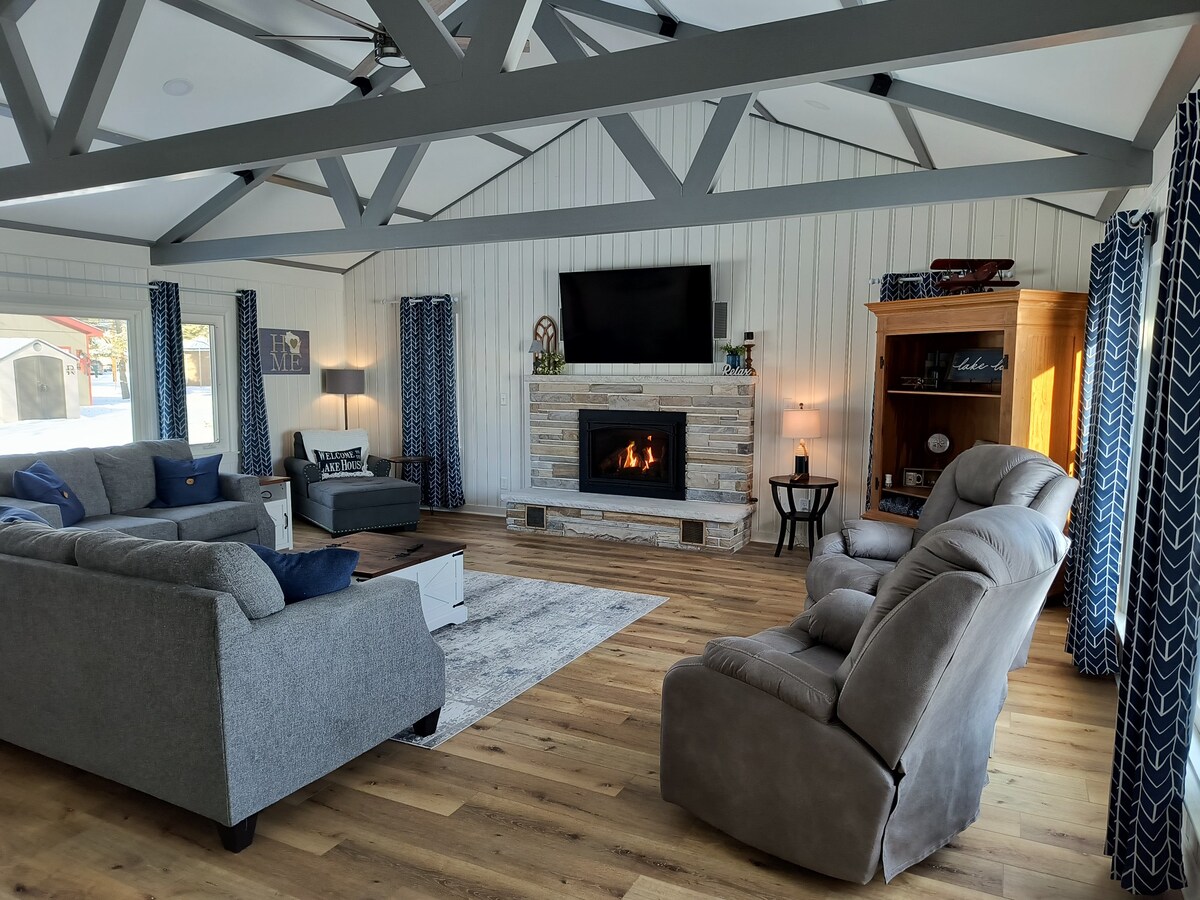 A spacious living room features a modern fireplace with a stone surround, flanked by large windows. Comfortable gray sofas and a reclining chair provide ample seating. Decorative curtains add texture, while a TV is mounted above the fireplace, enhancing the inviting environment.