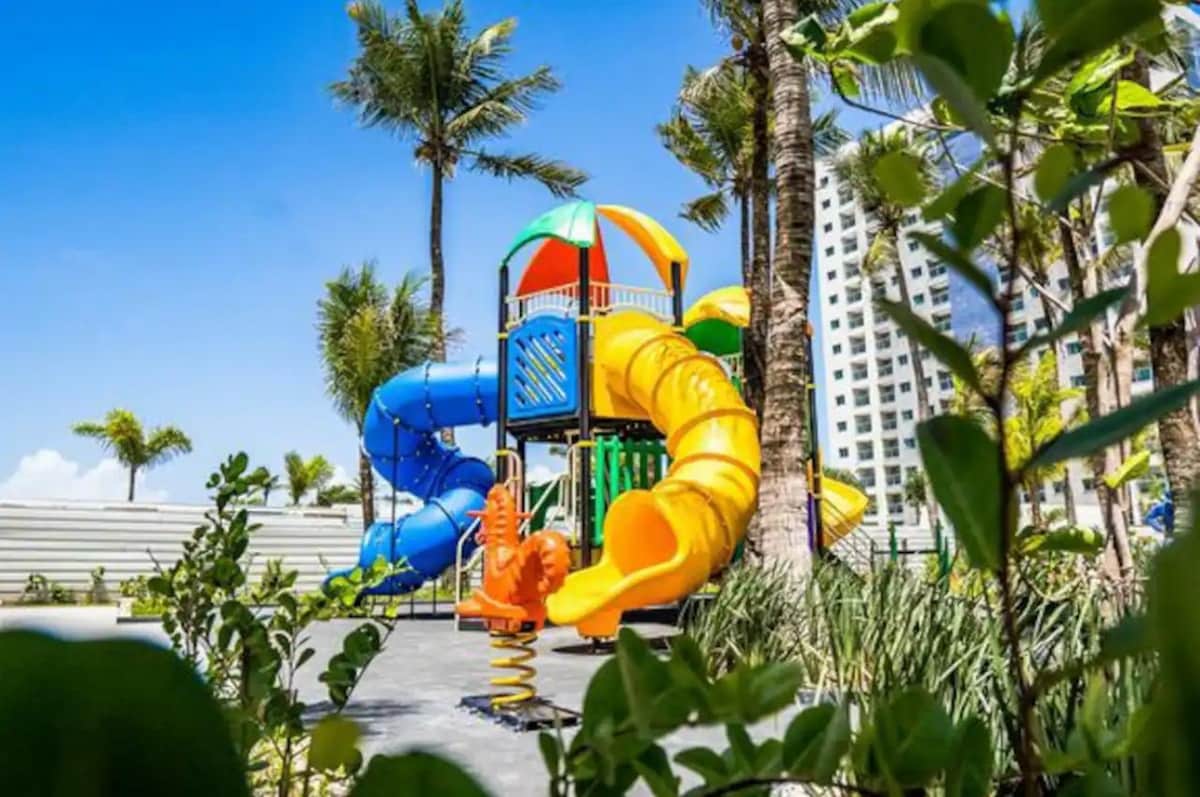 A vibrant playground designed for children is surrounded by palm trees, featuring colorful slides and climbing structures. The bright design includes a large yellow slide and a multi-level playhouse topped with a canopy. Lush greenery adds a natural touch to the playful environment.