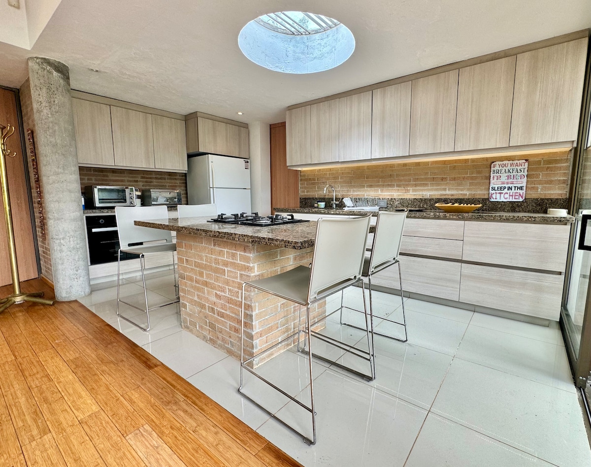 A modern kitchen features light wood cabinetry and a large island with a stone countertop. Stainless steel appliances include a refrigerator and an oven. Four chrome bar stools are arranged around the island, while natural light pours in from a circular skylight above.