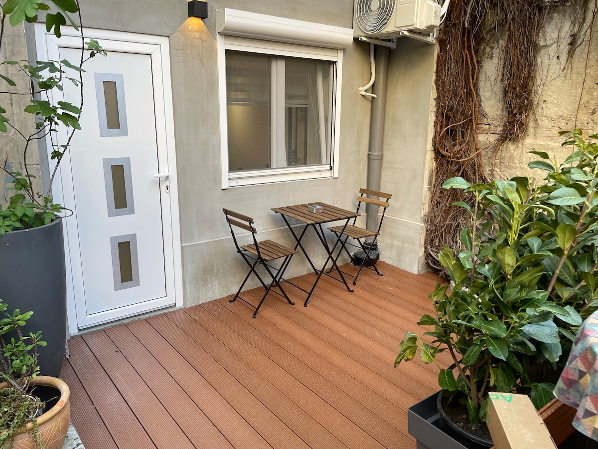 An outdoor terrace area features a small wooden table with two folding chairs, ideal for enjoying meals al fresco. Lush plants in pots surround the space, and a wall-mounted air conditioning unit is visible. Natural light filters through a nearby window.
