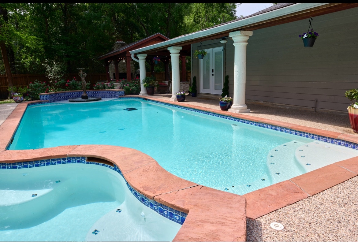 A sparkling pool is framed by a curved edge and surrounded by elegant columns. A relaxing spa is integrated beside the pool, with flower pots adding color. Lush greenery can be seen in the background, enhancing the tranquil outdoor setting.