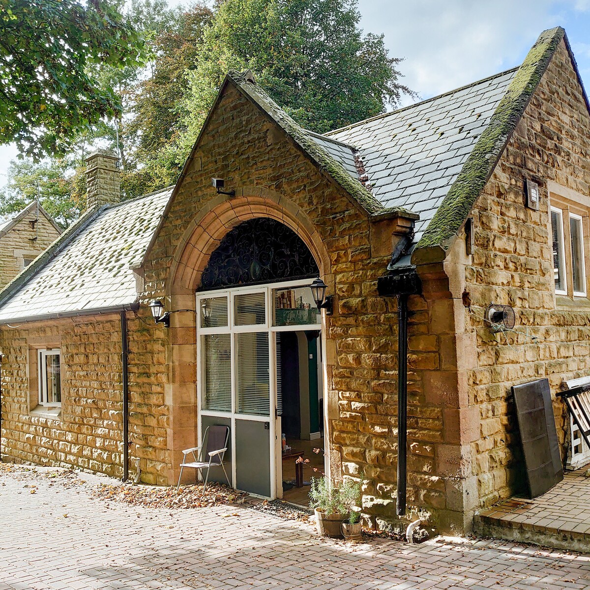 An exterior view of the unique outbuilding features traditional stone architecture and a steeply pitched roof. Large glass doors create a welcoming entrance, complemented by outdoor seating and landscaped details. Trees provide a natural backdrop, enhancing the serene atmosphere of the property.