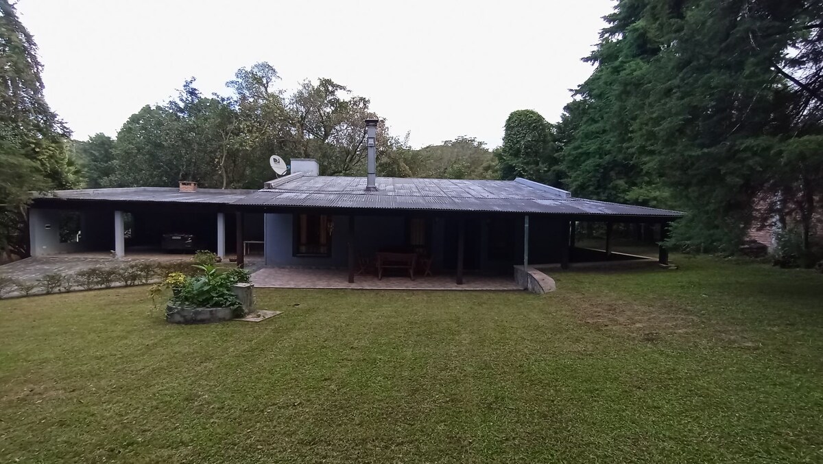 A spacious exterior of a house is surrounded by lush greenery, featuring a wide overhanging roof and a stone pathway. Outdoor seating is visible on a grassy area, while large trees provide shade and enhance the serene setting.