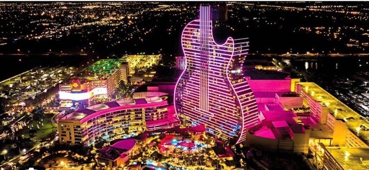 An aerial view reveals a vibrant hotel shaped like a guitar, illuminated with colorful lights. The surrounding area showcases a mix of attractions and amenities, creating a lively atmosphere. The city skyline enhances the backdrop, reflecting a blend of urban life and entertainment options.