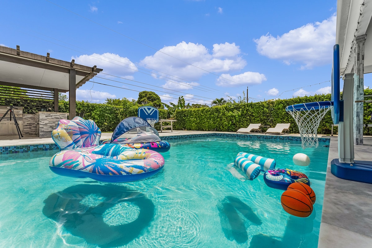 The heated pool is adorned with colorful floats, including lounge chairs and pool toys. A basketball hoop stands at the edge, ready for play. Surrounding greenery offers privacy, while the sky is bright with a few fluffy clouds.