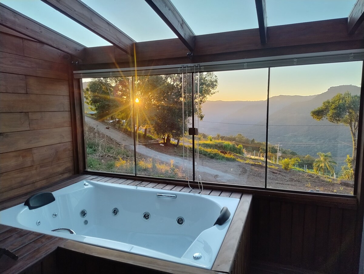 A spacious hot tub is positioned near large glass windows, offering views of a valley and sunset beyond. Natural light fills the area, accentuating wooden walls and the serene outdoor landscape.