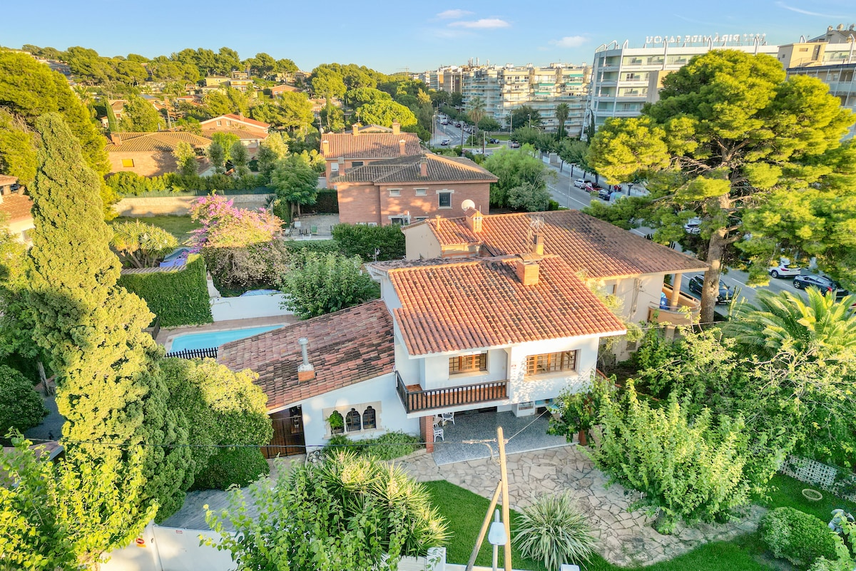 An aerial view captures the expansive villa with terracotta roofing, surrounded by lush greenery and a vibrant garden. The private pool and backyard area are visible, along with nearby buildings and trees that create a serene atmosphere.