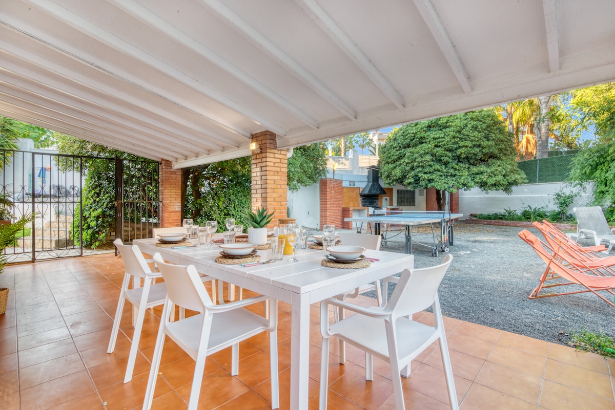 An outdoor dining area is positioned under a covered patio, featuring a white table set for six. The space is surrounded by greenery, with a ping pong table visible in the backyard. The flooring is tiled, providing a clean, open atmosphere.