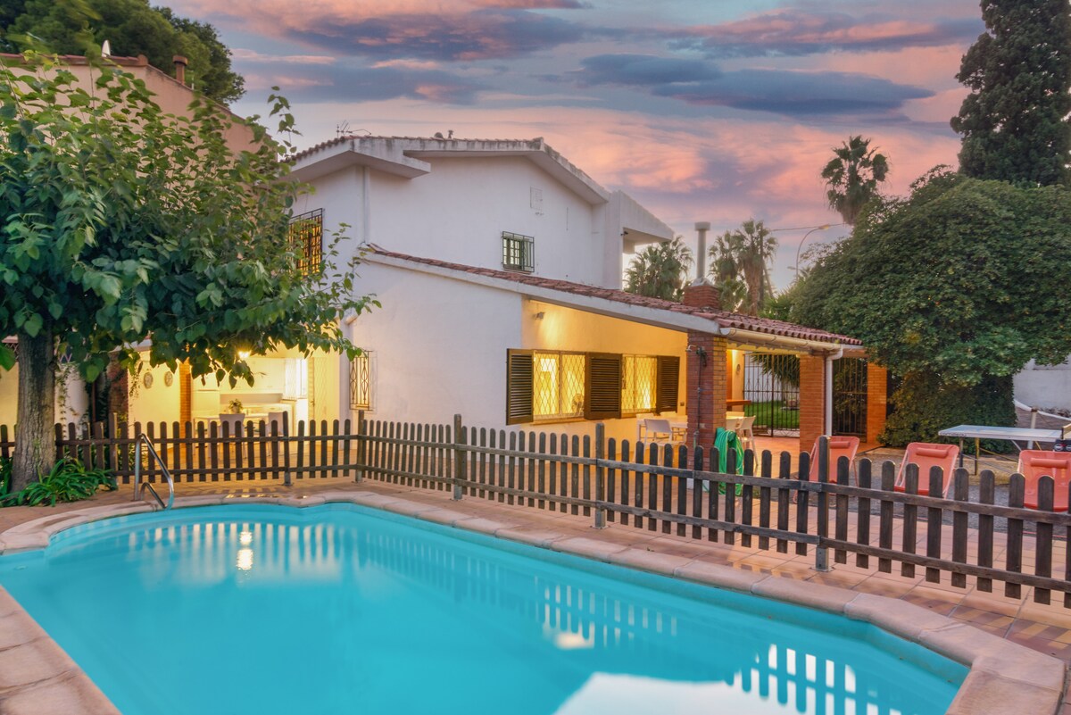 A private pool is framed by a wooden fence, reflecting the colors of a vibrant sunset. The villa features large windows with latticework and lush greenery surrounding the property, creating a serene outdoor space for relaxation. Chairs are placed near the pool, inviting leisure.