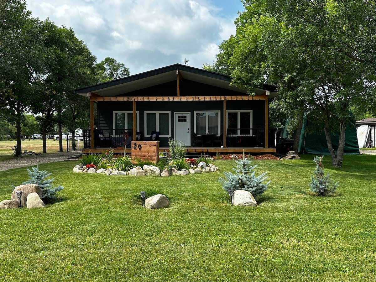 The exterior of the house features a covered front deck with seating and a landscaped garden. A well-maintained lawn surrounds the property, complemented by decorative stones and small trees. A serene, natural backdrop enhances the inviting appearance of the home.