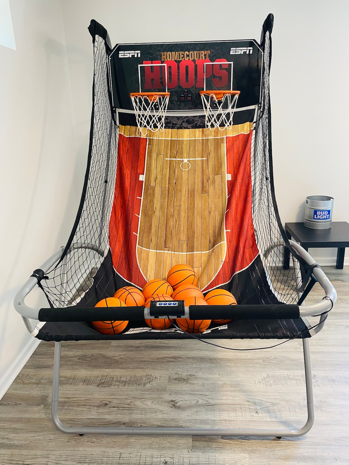 A basketball arcade game is positioned against a light-colored wall, featuring a colorful graphic of a basketball court. Several inflatable basketballs rest in front of the netted scoring area, ready for play. The flooring is a neutral-tone laminate, enhancing the recreational atmosphere.