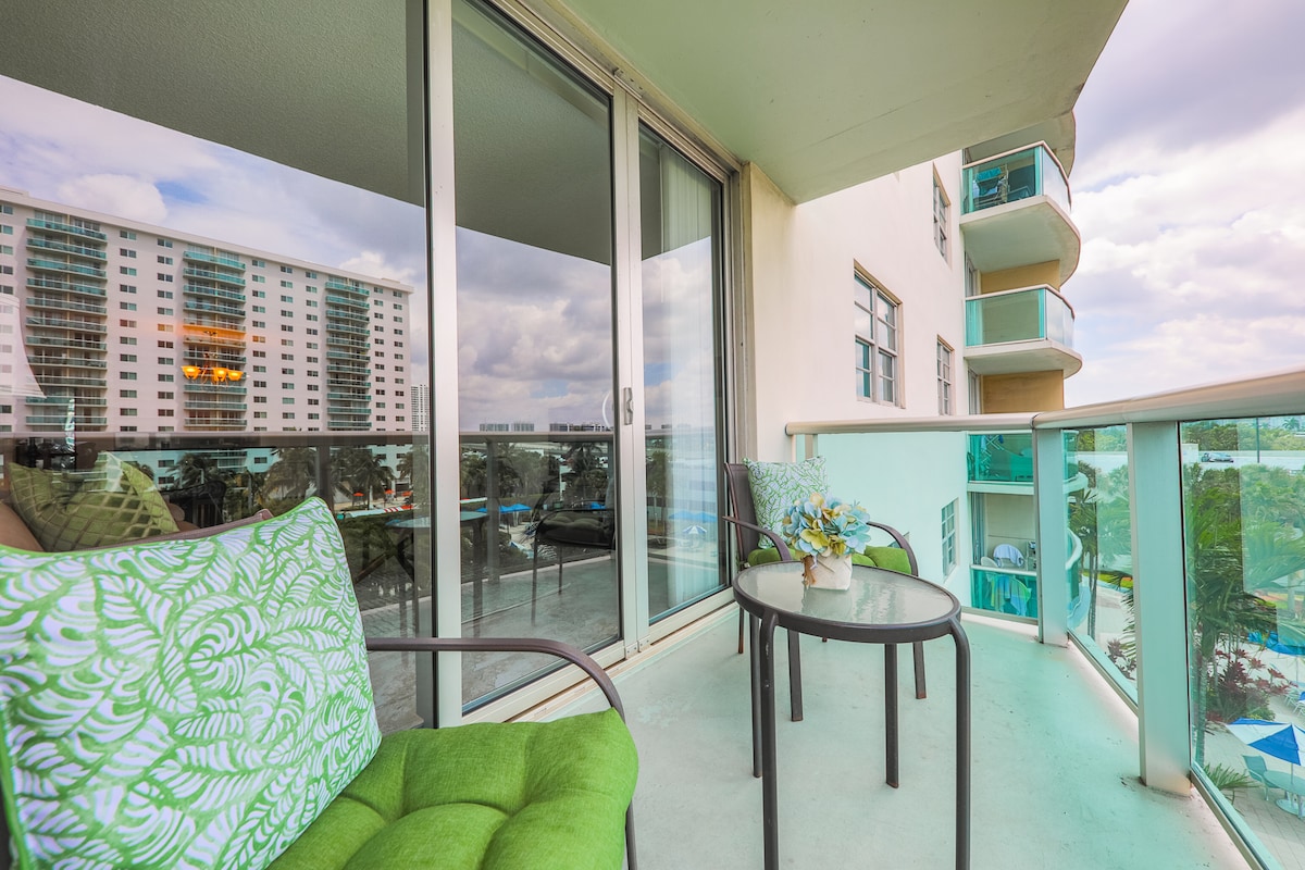 A private balcony features two chairs, one adorned with a green cushion, and a small table. The glass railing offers unobstructed views of the surroundings. Large sliding doors provide access to the interior, while a bright sky and distant buildings are visible in the background.