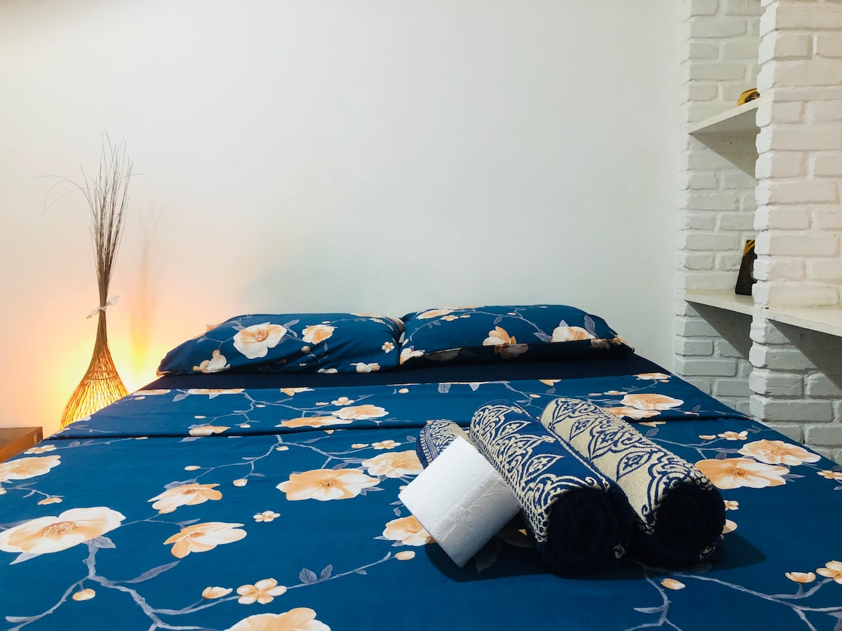 A cozy sleeping area features a spacious bed dressed in floral blue bedding. Soft pillows are neatly arranged, while rolled towels are placed at the foot of the bed. A warm light from a decorative lamp enhances the tranquil ambiance of the room.