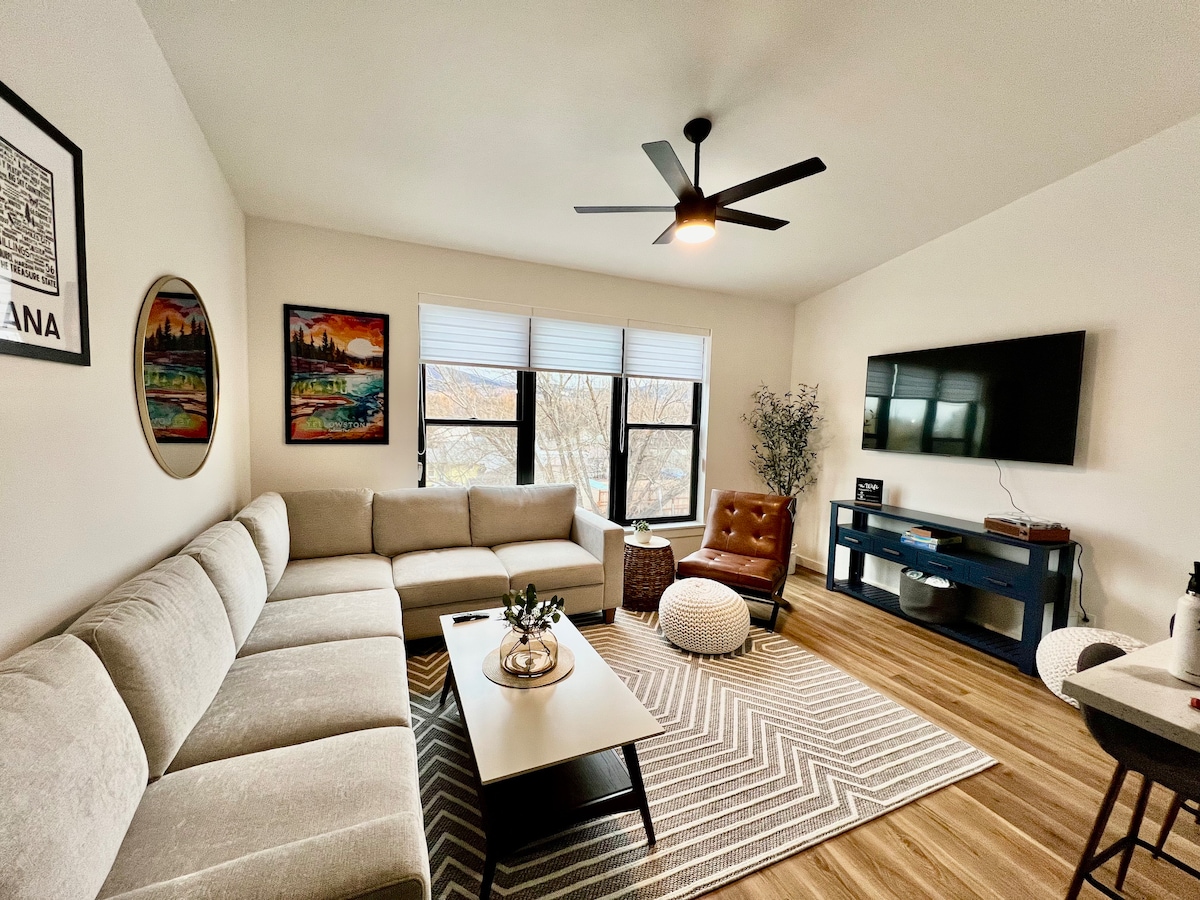 A spacious living area is furnished with a large sectional couch and offers seating on an accent chair. Natural light enters through multiple windows, and a wall-mounted TV is positioned for entertainment. A textured area rug adds warmth to the polished wooden floor.