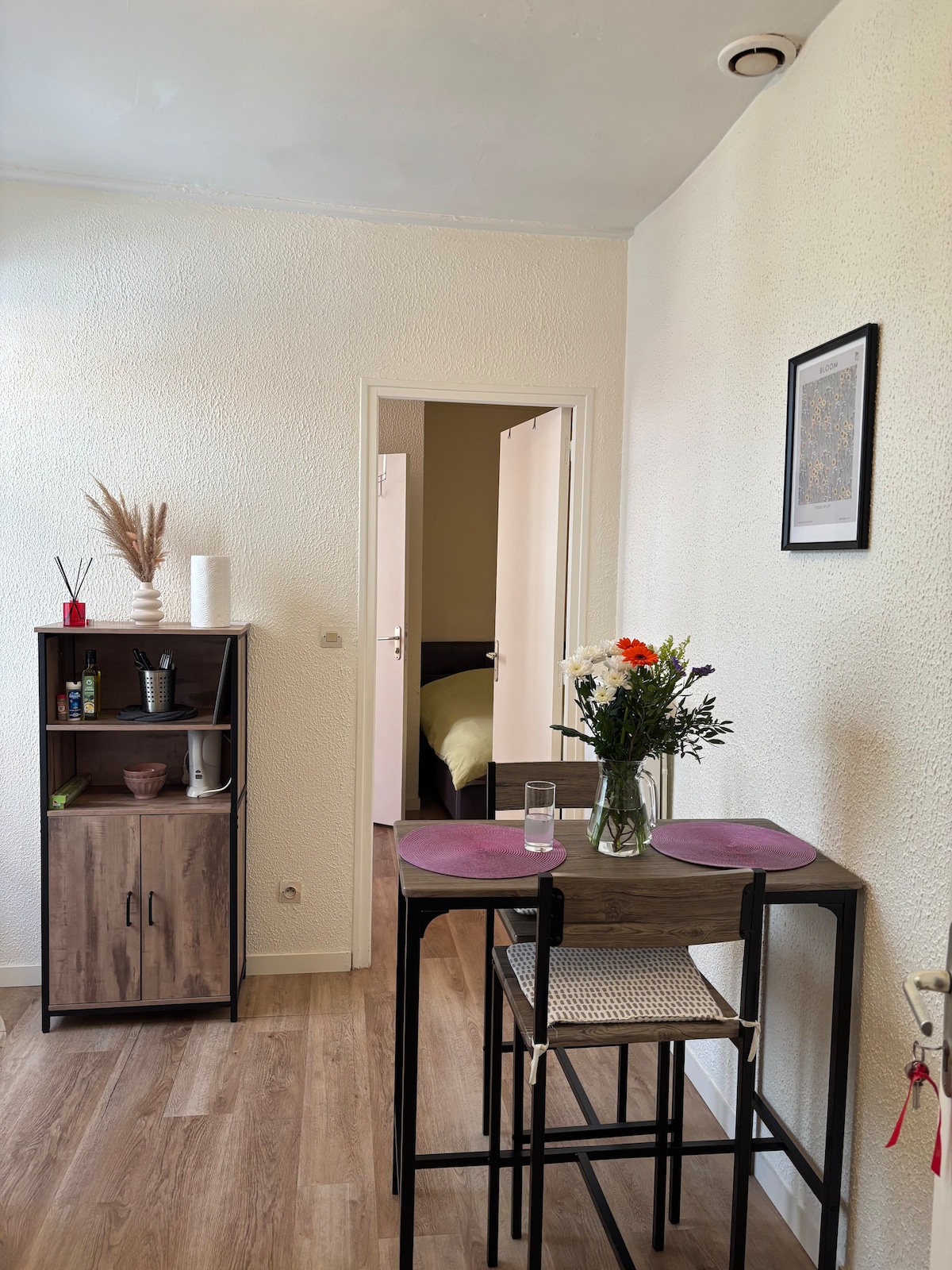 A compact dining area is presented, featuring a small table with two chairs adorned with light purple placemats. A cabinet is situated nearby, displaying decorative items. A light-filled doorway leads to a bedroom in the background, indicating a functional layout.