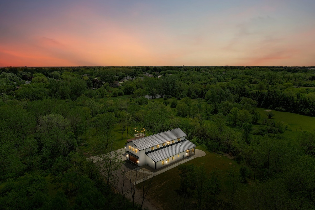 The exterior view of the barnhouse reveals a tranquil setting, surrounded by lush greenery under a soft twilight sky. The structure showcases large windows that reflect the warm glow of interior lights, suggesting a welcoming atmosphere for guests.
