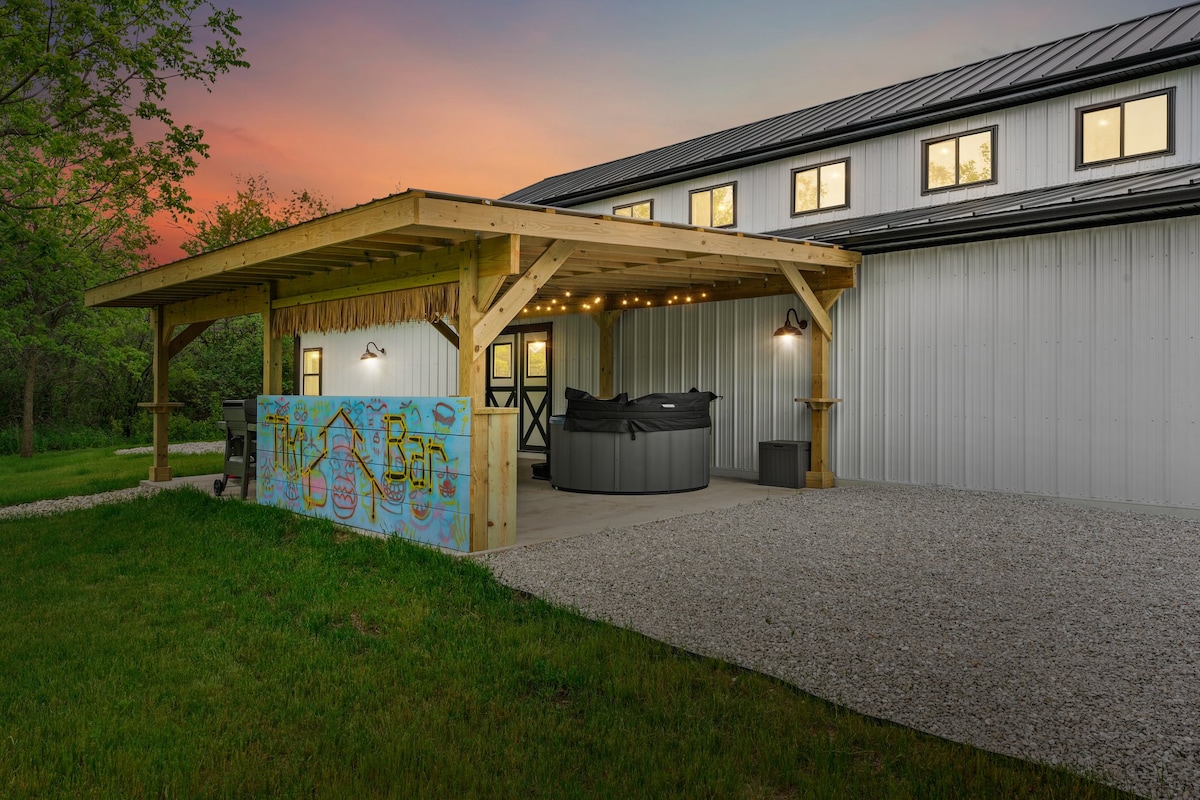 An outdoor lounge area features a covered pergola with an illuminated Tiki Bar and a hot tub. The gravel pathway leads to the entrance of the barn, surrounded by lush green grass and trees, creating a serene atmosphere at sunset.