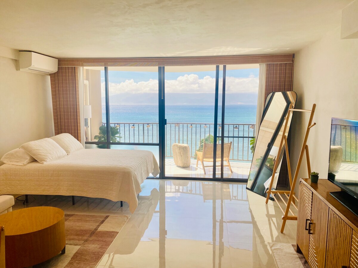A bright studio is presented with large glass doors offering expansive ocean views. A queen bed is placed in the corner, alongside a sitting area with a chair and a small table. A mirror and a TV stand enhance the spacious layout, with natural light flooding the room.