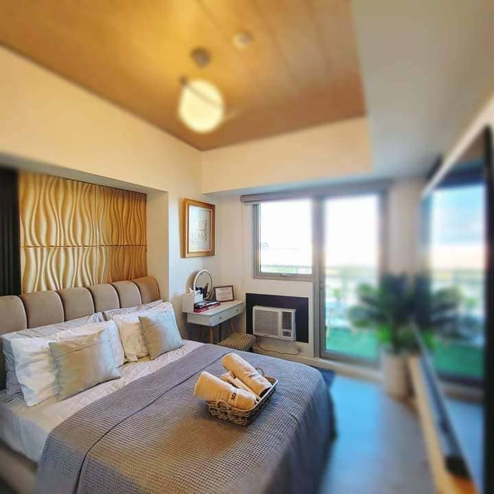 A comfortable bedroom features a spacious bed with neatly arranged pillows and a textured headboard. A coffee table and a small seating area are present, while large windows allow natural light to fill the room. Air conditioning and a small plant add to the ambiance.