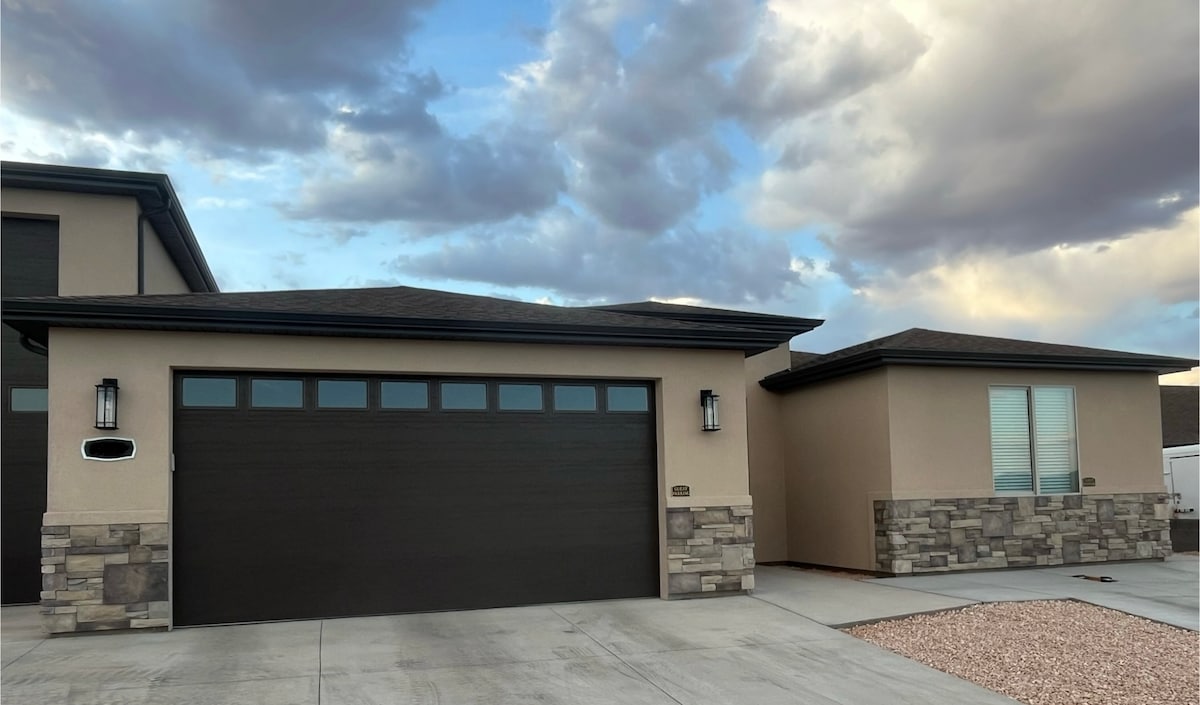 A modern casita exterior is shown, featuring a combination of stucco and stone accents. A large garage door is prominently displayed, framed by stylish wall sconces. The driveway is paved with a smooth finish, and a gravel landscape decorates the front area under a partly cloudy sky.