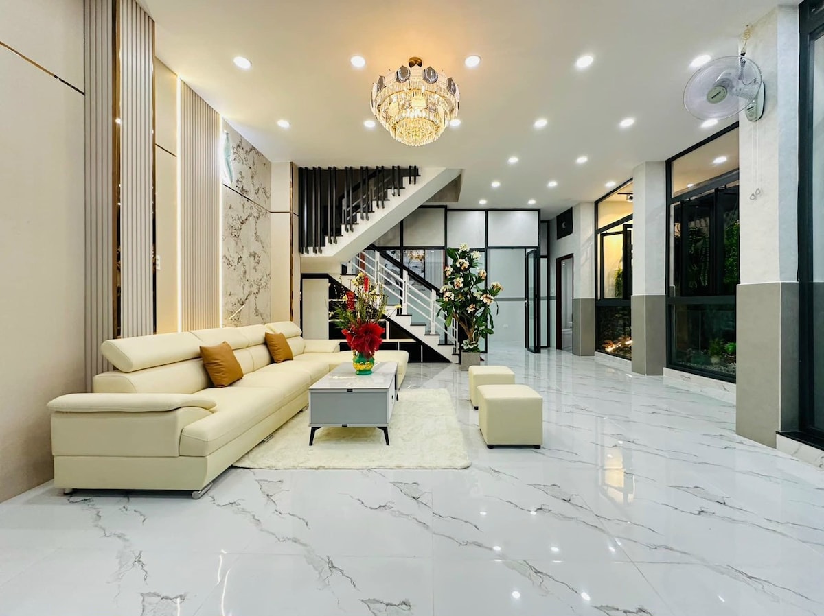 A spacious living area features a light-colored sectional sofa with decorative cushions. A glass coffee table is positioned on a plush rug, surrounded by vibrant floral arrangements. The room is illuminated by recessed lighting and a modern chandelier, with a staircase visible in the background.
