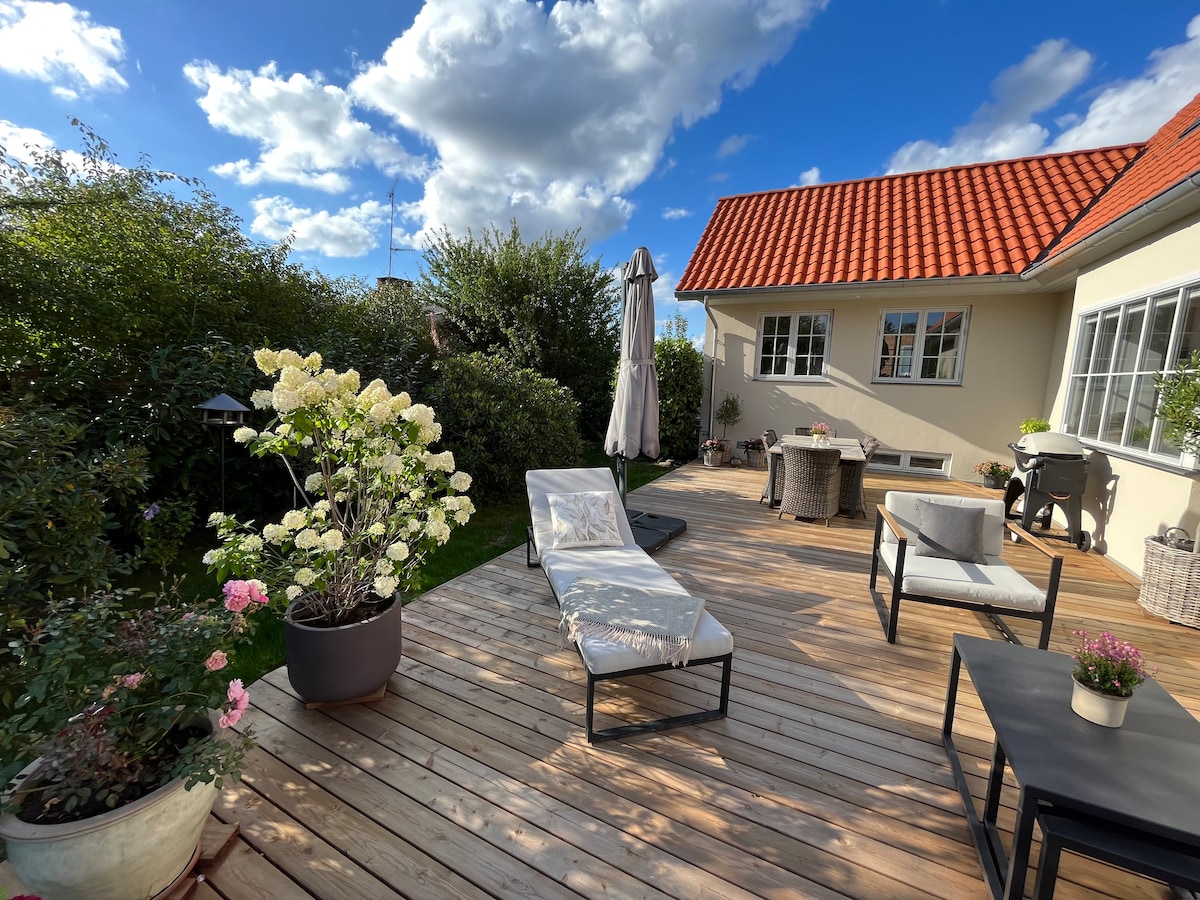 A spacious wooden terrace is surrounded by lush greenery, featuring comfortable lounge chairs and a parasol. Potted flowers add color, while a seating area with a table and chairs is visible, inviting relaxation and outdoor dining.