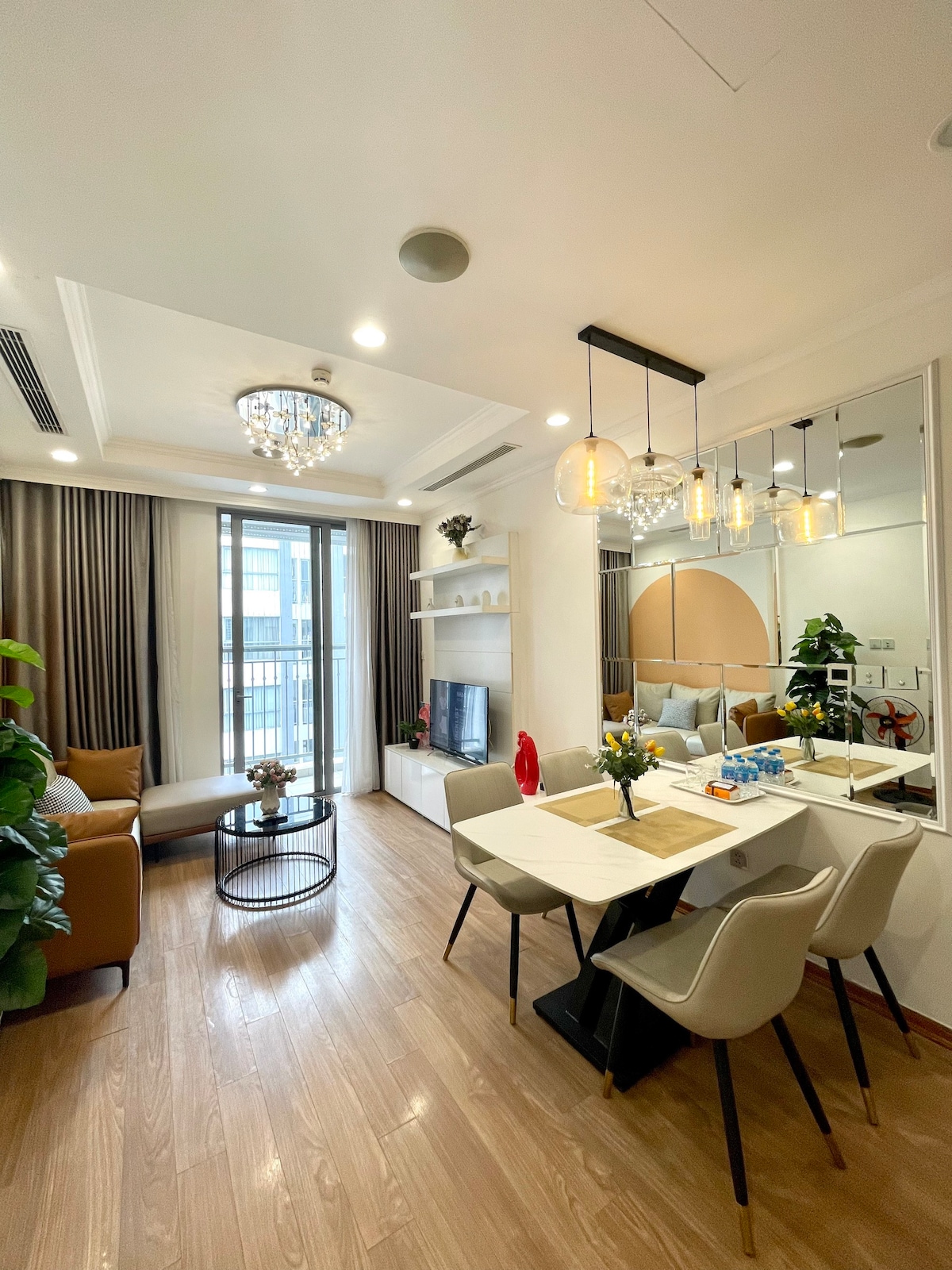 A modern living area features a cozy brown sofa and a round coffee table accompanied by a decorative plant. A dining table for four is illuminated by pendant lights, with a mirror enhancing the spacious feel. Large windows invite natural light, offering views of the outside.