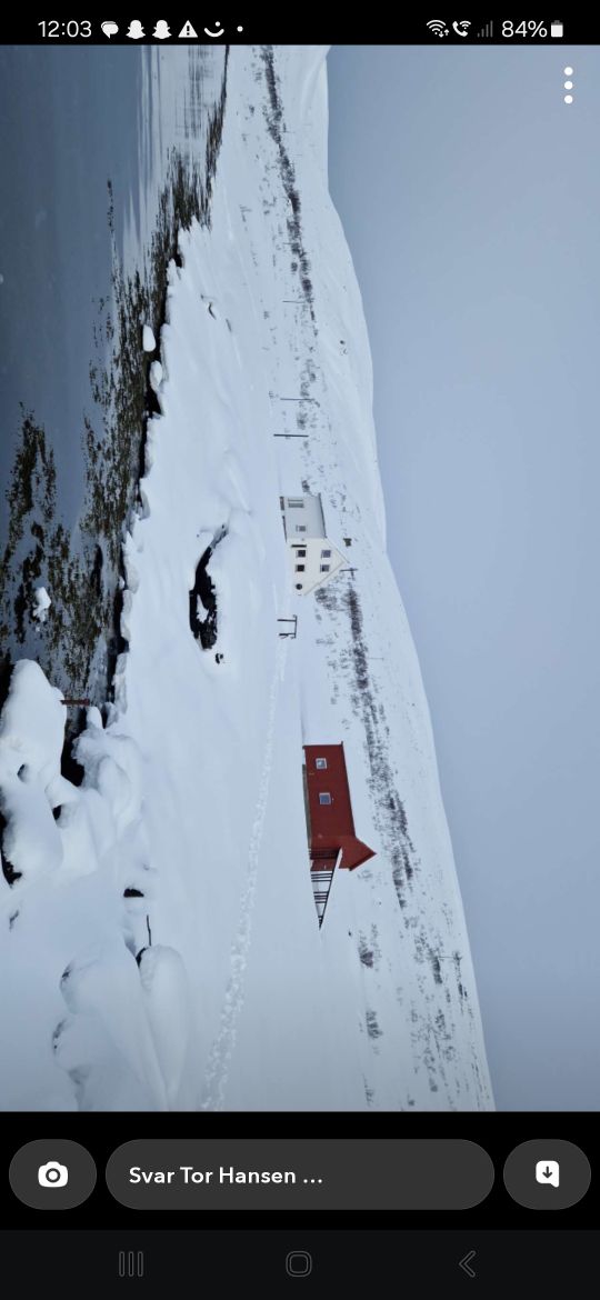 A serene wintry landscape is depicted, showcasing a red building contrasting against the white snow. Rolling hills are covered with snow, extending into a calm body of water. Footprints lead through the snow, guiding towards the structures in the distance.