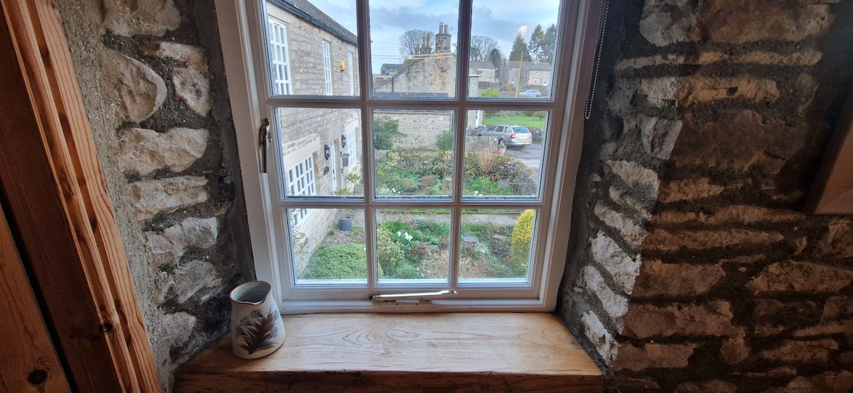 A window framed by rustic stone walls provides a view of a landscaped garden and surrounding buildings. A small ceramic mug rests on the wooden windowsill. Outside, greenery and vibrant blooms add a touch of color to the serene setting.