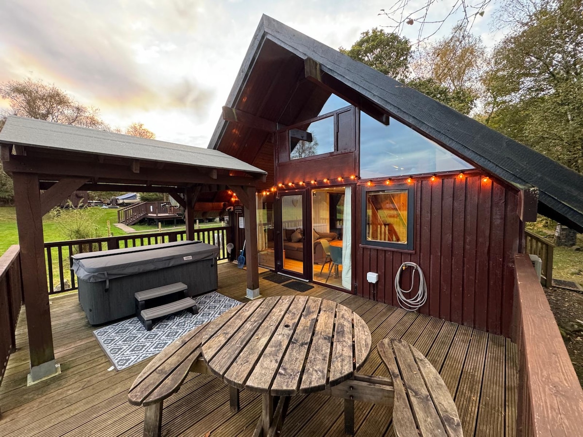 An outdoor deck features a large wooden table surrounded by benches, illuminated by string lights. An electric hot tub is positioned nearby, and floor-to-ceiling glass doors provide access to indoor spaces, framed by natural surroundings.