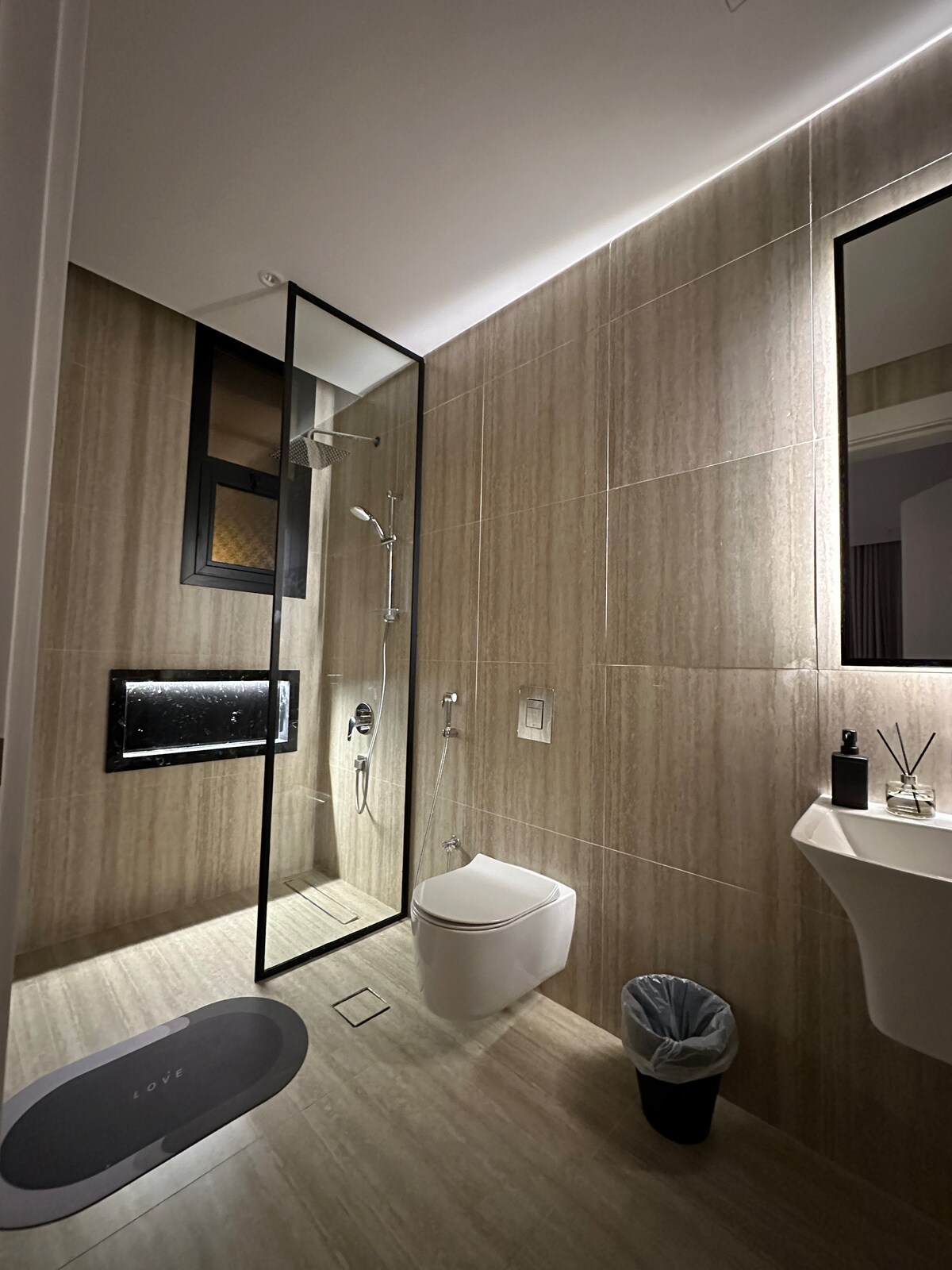 A modern bathroom is depicted, featuring a spacious shower enclosed by glass panels. Beige tiled walls complement the sleek black accents. A white toilet and sink are positioned to create a functional layout. Natural light filters through a small window, enhancing the serene environment.