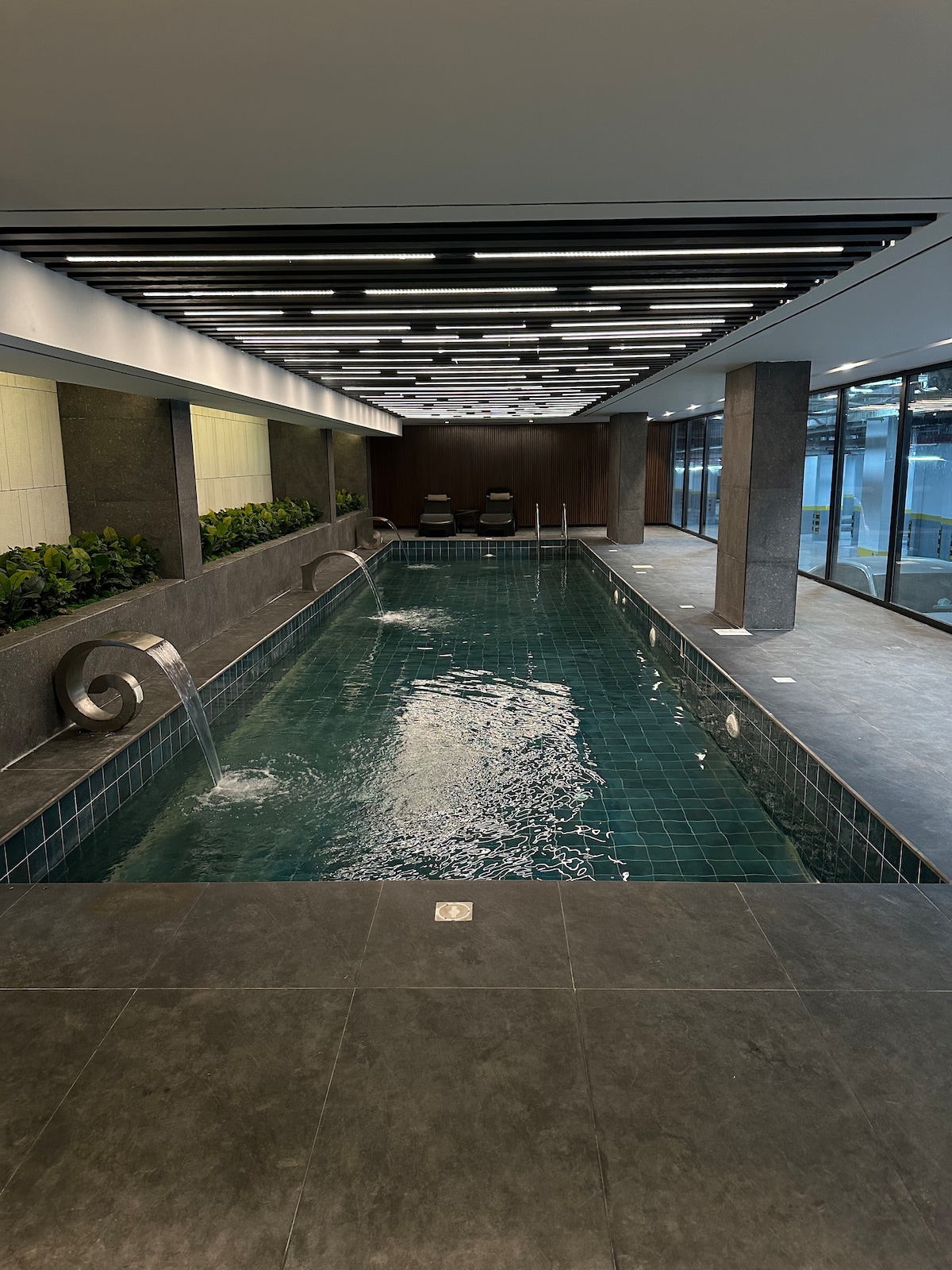 An indoor swimming pool is surrounded by sleek stone walls and modern lighting. Water features create gentle cascades, while comfortable loungers are positioned beside the pool. Large windows provide views of the exterior, enhancing the tranquil atmosphere.