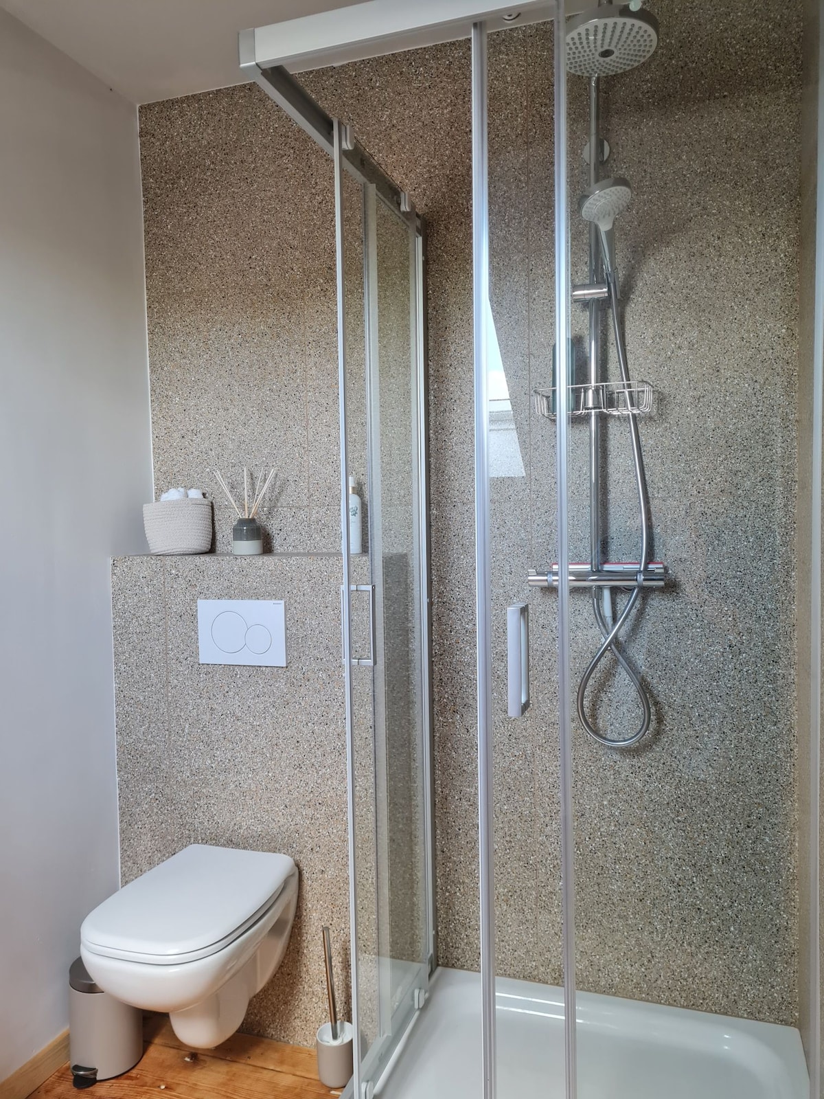 The bathroom features a glass shower enclosure with a handheld showerhead and wall-mounted accessories. A modern toilet is positioned alongside a textured wall. Neutral tones are complemented by a small, organized shelf with toiletries and decorative items.