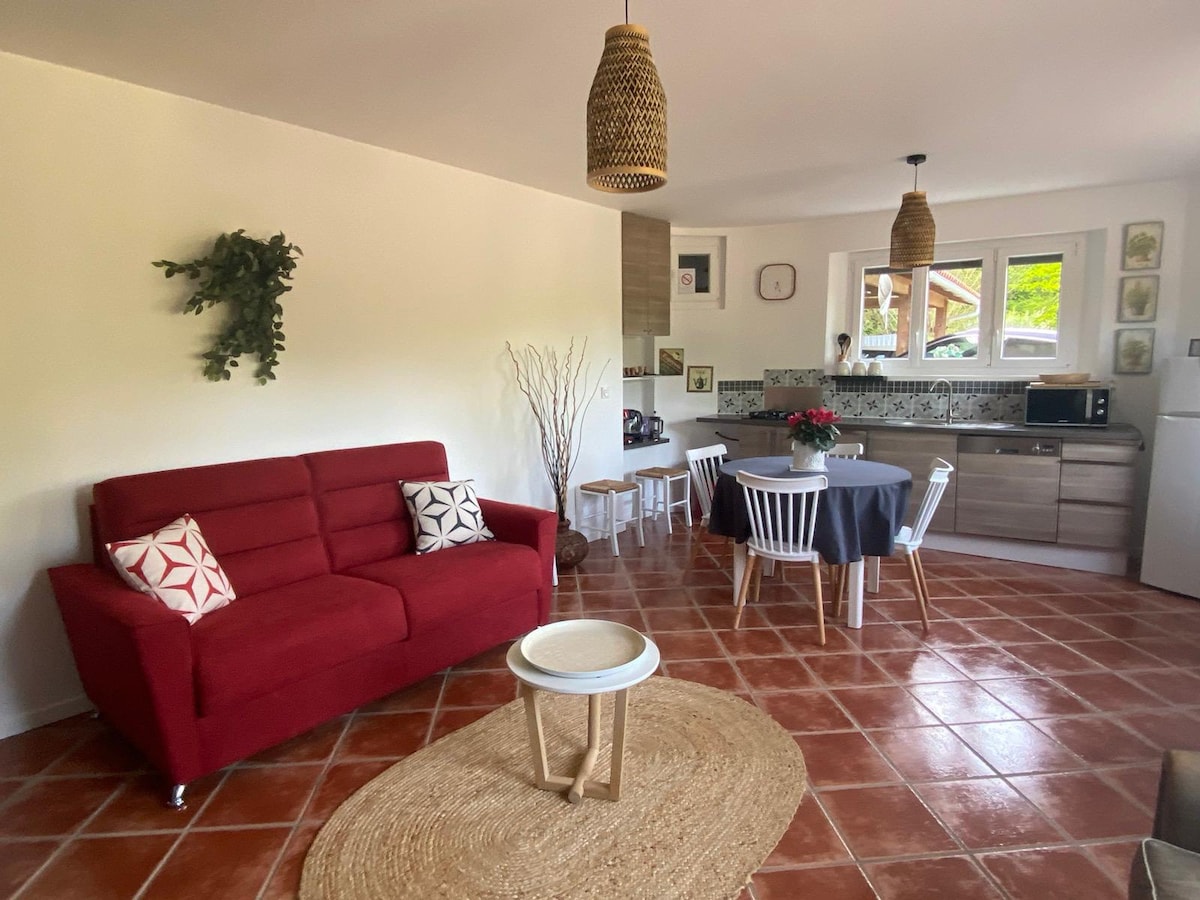 A cozy living space features a red sofa with decorative cushions. A round dining table is surrounded by white chairs, while a kitchen area is equipped with modern appliances. Light enters through the window, illuminating the terracotta tiled floor and the natural textures of decor.