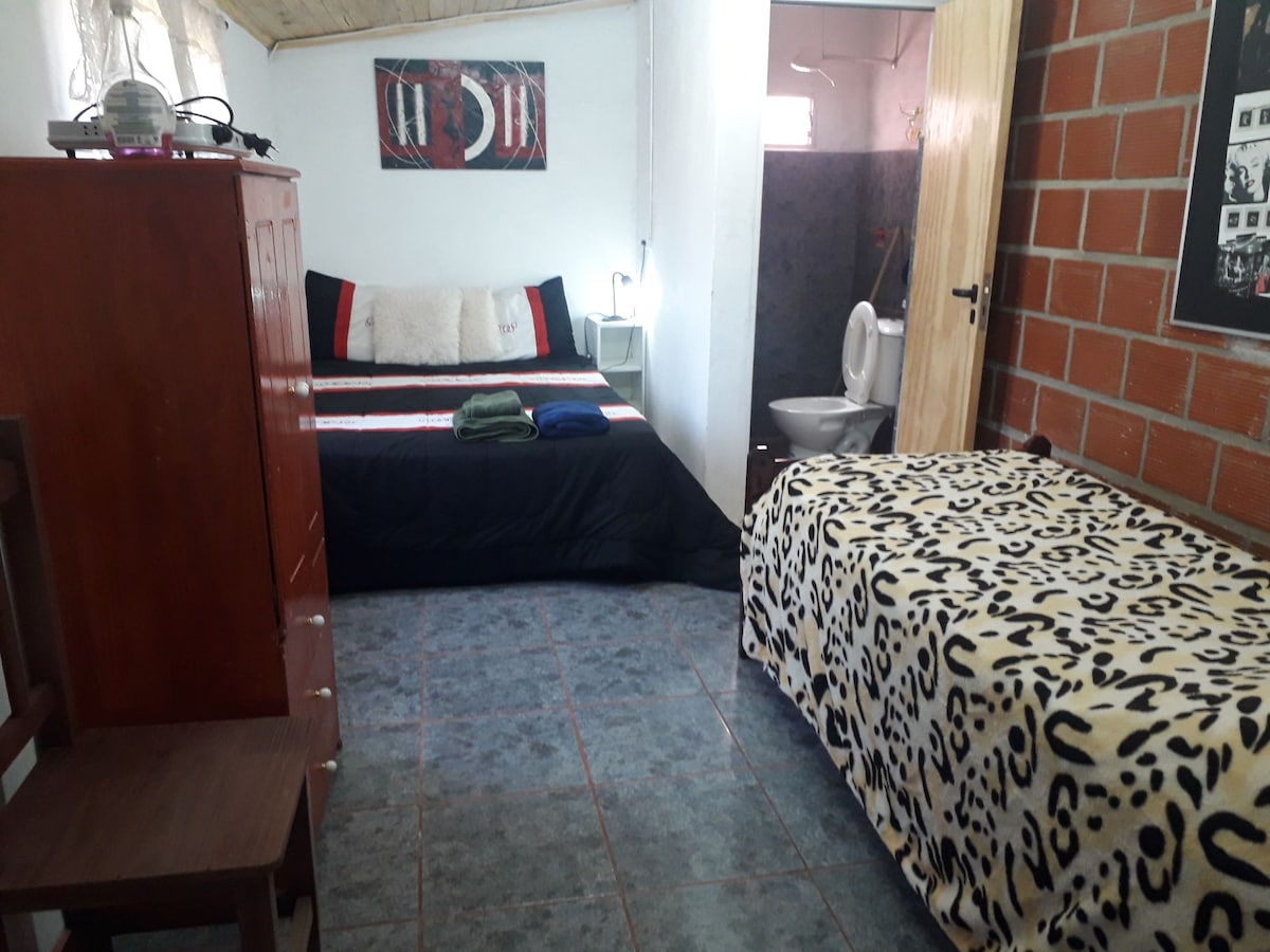 A cozy room features a spacious bed with neutral linens, complemented by a contrasting black cover. A single bed is dressed in a patterned cover, while a wooden chair and a chest of drawers occupy the corner. A private bathroom is accessible in the background.