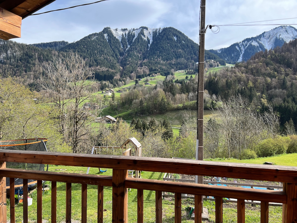 A scenic view from the balcony reveals lush green hills and majestic mountains. The landscape is dotted with trees and rustic farmhouses, creating an atmosphere of tranquility. Soft sunlight illuminates the surroundings, highlighting the natural beauty of the area.