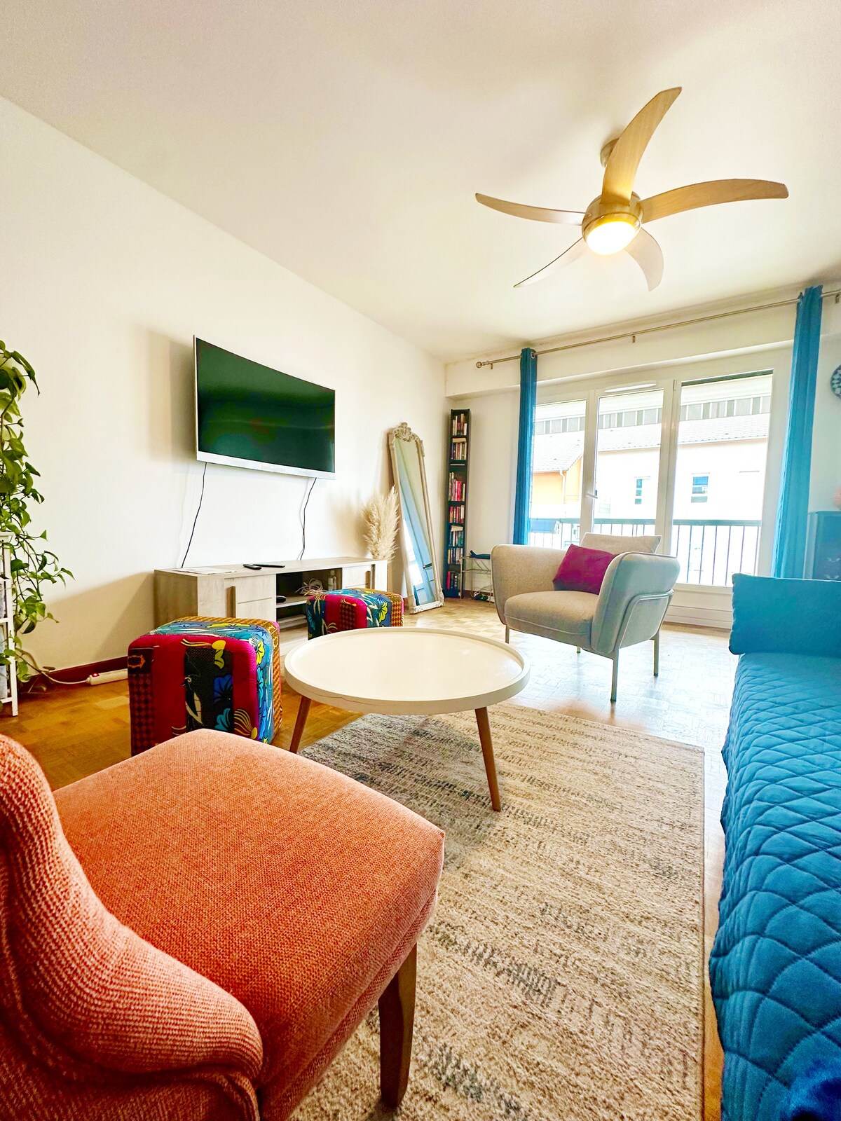 A cozy living space features a television mounted on a wall, with comfortable seating arrangements including two accent chairs and a blue couch. Natural light fills the area through large windows, creating a bright atmosphere. Decorative plants add a touch of greenery.